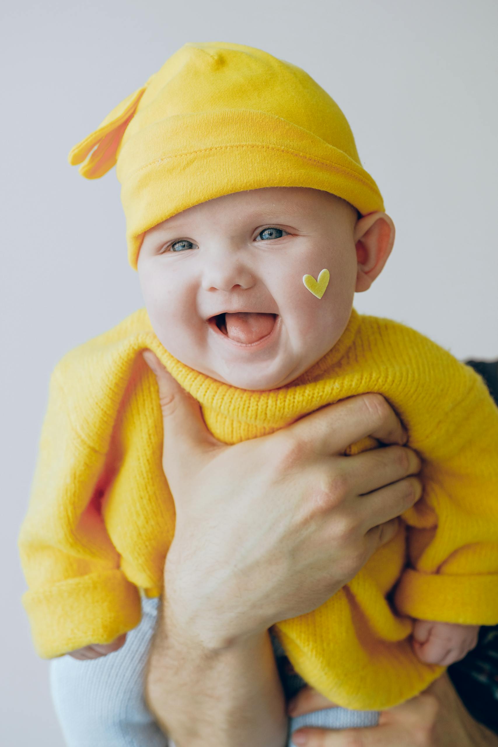 Adorable baby wearing a yellow beanie with a heart sticker, smiling joyfully indoors.