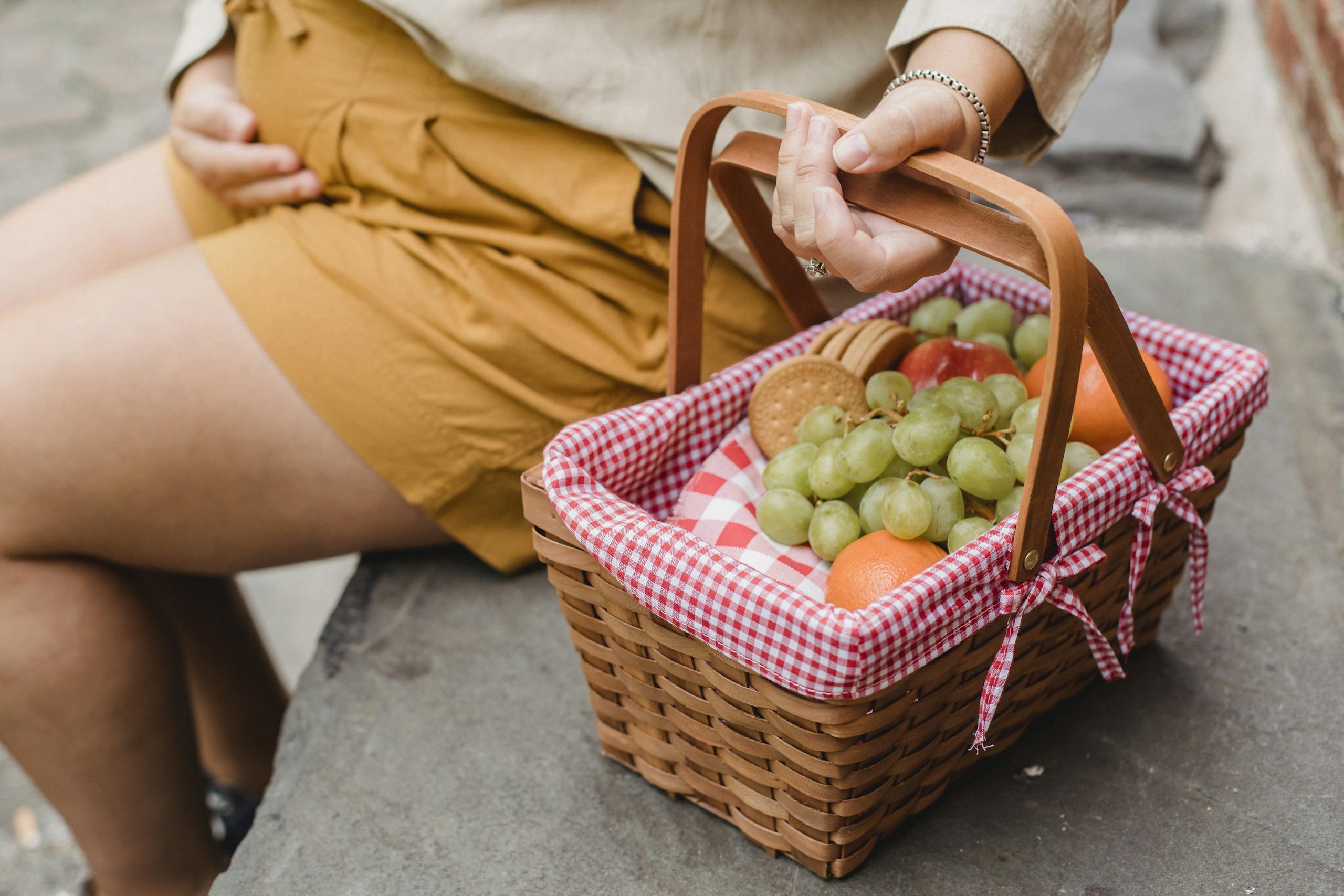 Crop anonymous pregnant female in casual outfit with basket full of fresh ripe fruits and tasty cookies prepared for picnic