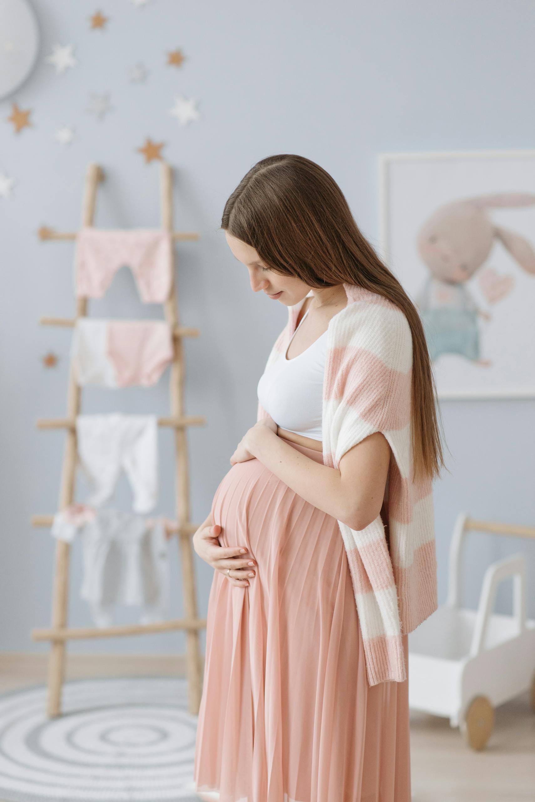 Pregnant woman in pink skirt caressing baby bump in serene nursery setting.