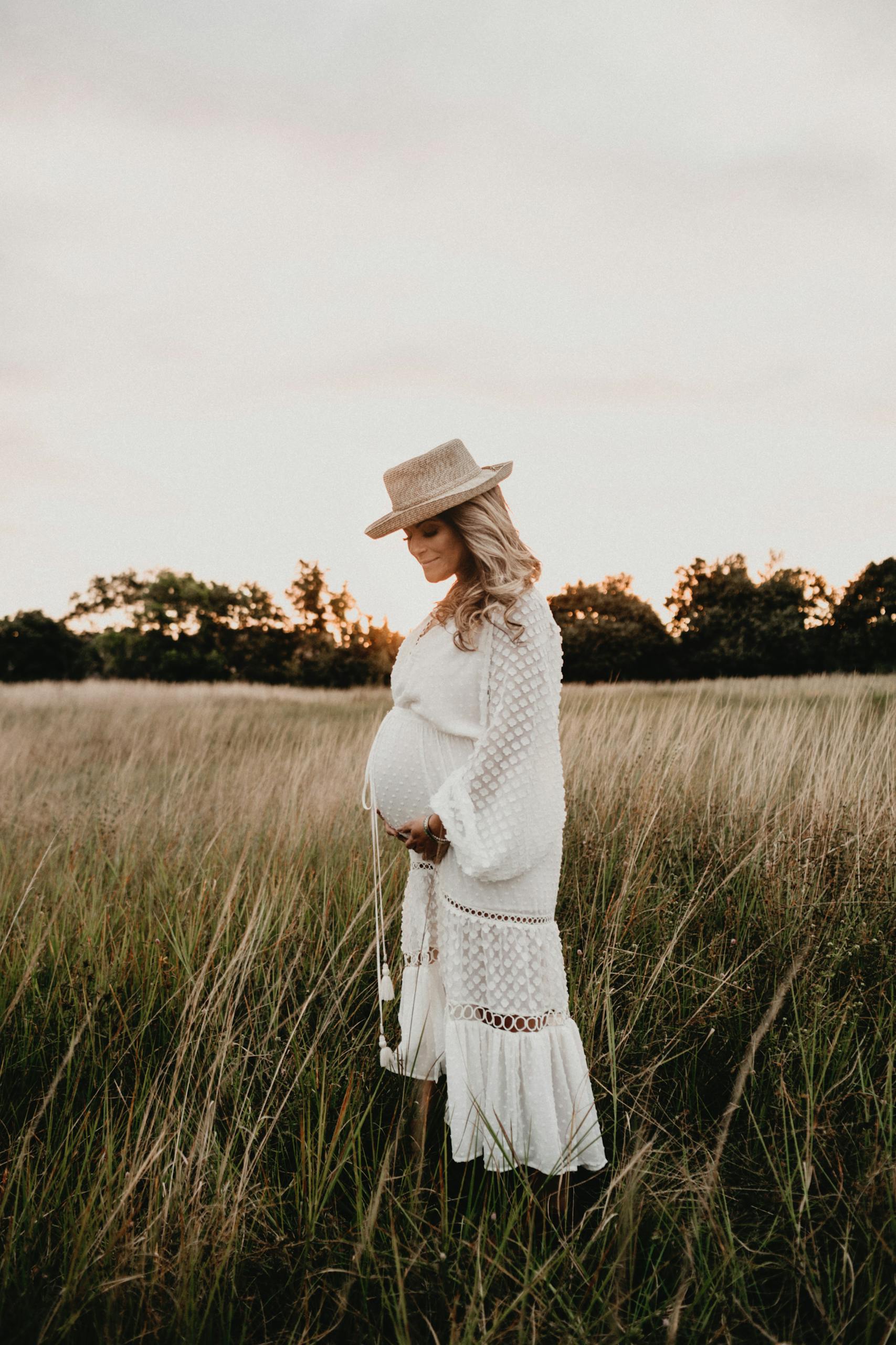 Stylish pregnant woman in a white dress and hat standing in a grassy field during sunset.